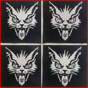 May include: Four square black fabric patches featuring a white graphic of a snarling cat head. The cat's eyes contain the text 'THEY THEM', 'SHE THEY', 'HE HIM', and 'HE THEY'. An anarchy symbol with a female gender sign is on the tongue.