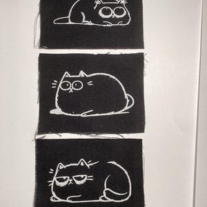 May include: Three rectangular black fabric patches, each featuring a white line-art illustration of a cat. The top cat has wide eyes, the middle cat is round and plump, and the bottom cat wears glasses with a grumpy expression.