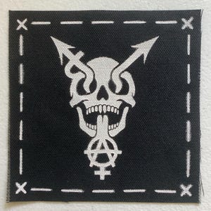 May include: A square black fabric patch featuring a white screen-printed graphic of a skull with two upward-pointing arrows emerging from the eye sockets and an anarchy symbol combined with a female gender sign extending from the open mouth.