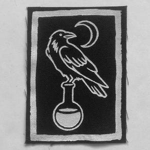 May include: A rectangular black fabric patch featuring a white screen-printed design of a raven perched atop a round-bottomed laboratory flask. Above the bird is a crescent moon. The entire graphic is enclosed within a thick white rectangular border.