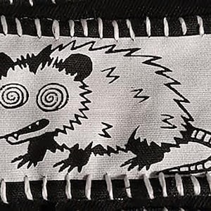 May include: A black and white patch featuring a cartoon opossum with spiral eyes and a zig-zag tail. The opossum is stitched onto a black fabric with white stitching around the edges. The design is simple and bold.