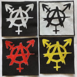 May include: Four square patches with a symbol combining the anarchy symbol, a male symbol, and a female symbol. The patches are black, white, red, and yellow. The symbols are white, black, red, and yellow, respectively.