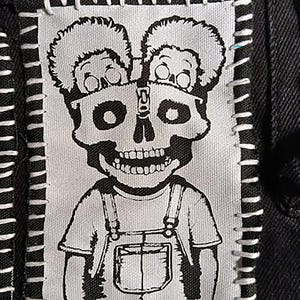 May include: A black and white patch featuring a cartoon skull with two smaller skulls above it. The skull is wearing overalls. The patch is sewn onto a black fabric with white stitching around the edges.