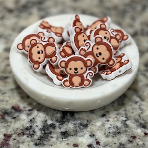May include: A collection of brown and tan monkey-shaped charms with white outlines, resting in a white marble bowl. The charms have a cartoon-like design, featuring a smiling face and a curved tail. The bowl sits on a speckled gray surface.