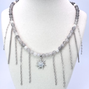 May include: A necklace featuring a strand of smooth grey beads interspersed with textured silver-toned spacer beads. Multiple silver-toned metal chains hang vertically from the bead strand, with a central sun-shaped pendant featuring clear stones.