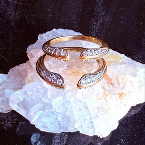 May include: A gold-toned ring with the name "CAMILLE" engraved on the band. The ring features a split design with two curved sections, each adorned with small, sparkling stones. The ring is displayed on a white crystal surface.
