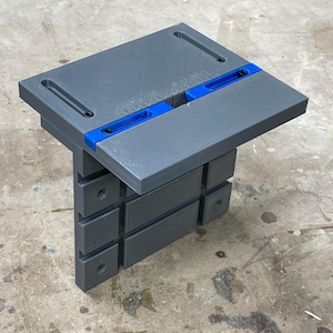 Op de afbeelding: Een grijs en blauw houtbewerkingsgereedschap met een rechthoekige bovenkant en een reeks gestapelde rechthoekige steunen. De bovenkant heeft een blauw accent en meetmarkeringen. De tool is ontworpen voor precisie en wordt waarschijnlijk gebruikt voor het snijden of vormen van hout.