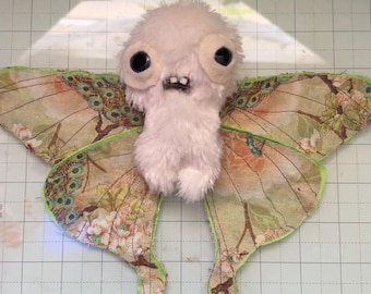 May include: A handmade plush creature with a fluffy white body, large black eyes, and a small mouth. It has large wings with a floral and peacock feather pattern, outlined in green. The creature is on a grid background.