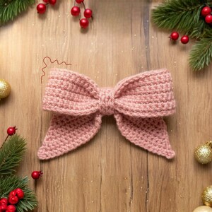 May include: A handmade, light pink crochet bow. The bow is centered on a wooden surface, surrounded by Christmas decorations, including red berries, pine branches, and gold ornaments. The bow's texture is clearly visible, highlighting the craftsmanship.