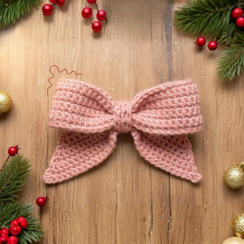 May include: A handmade, light pink crochet bow. The bow is centered on a wooden surface, surrounded by Christmas decorations, including red berries, pine branches, and gold ornaments. The bow's texture is clearly visible, highlighting the craftsmanship.