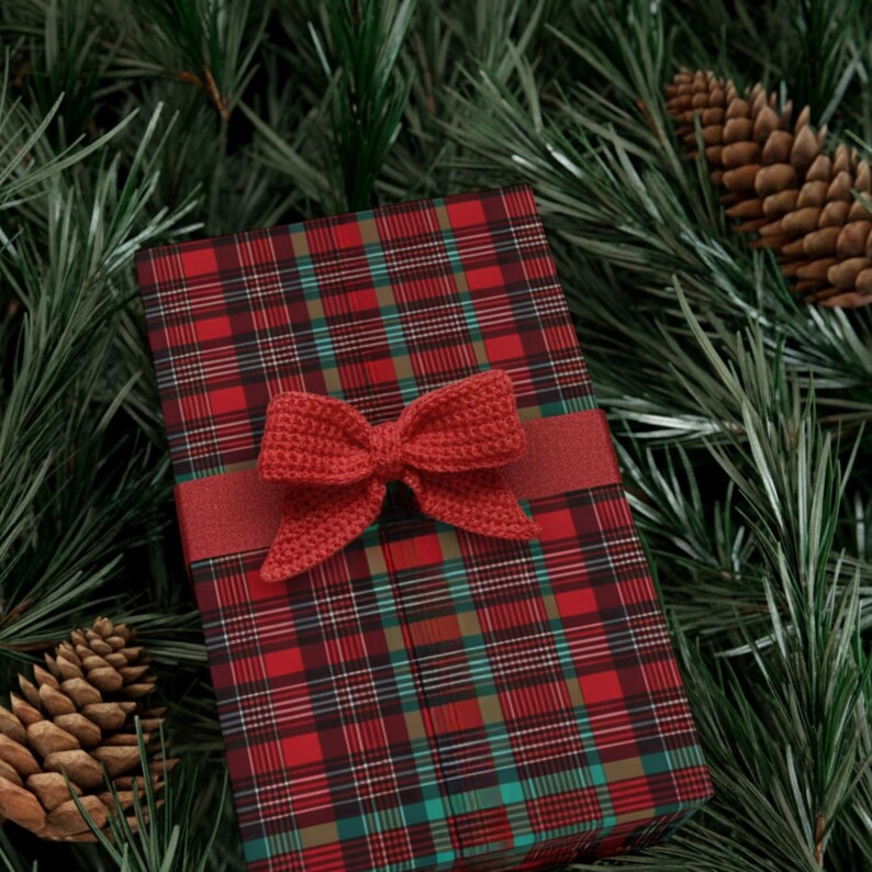 May include: A wrapped gift box with a red and green plaid pattern, adorned with a red crocheted bow and ribbon. The gift is surrounded by pine needles and pine cones, creating a festive holiday theme.