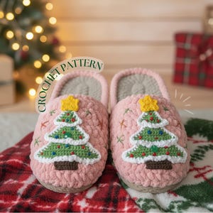 May include: A pair of pink crochet slippers with a Christmas tree design. The slippers feature a green tree with red and blue ornaments, a yellow star, and the text "CROCHET PATTERN". The slippers are on a red and green plaid blanket.