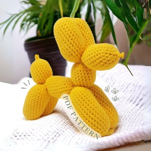 Op de afbeelding: Een gehaakt geel ballondier in de vorm van een hond. De gehaakte hond zit op een wit deken met de woorden "PDF PATTERN" erop.