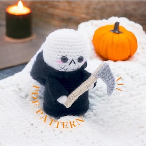 Op de afbeelding: Een gehaakte amigurumi Grim Reaper die een zeis vasthoudt. De Grim Reaper is wit met een zwarte mantel en heeft een schattig, cartoonachtig gezicht. De zeis is grijs en heeft een bruine steel. De Grim Reaper zit op een wit, pluche oppervlak. De tekst "PDF PATTERN" is zichtbaar in de afbeelding.