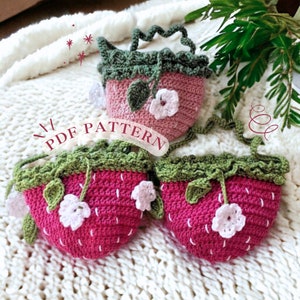 Op de afbeelding: Drie gehaakte aardbei-vormige tassen. De tassen zijn voornamelijk rood met groene toppen en witte bloemaccenten. De tekst "PDF PATTERN" is zichtbaar. De tassen hebben een handvat en een riem. De tassen liggen op een wit, getextureerd oppervlak.
