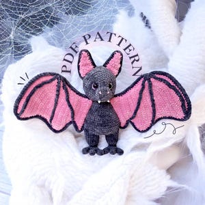May include: A crocheted bat amigurumi toy with pink wings and a gray body. The toy is sitting on a white fuzzy surface. The text "PDF PATTERN" is written in a circle around the toy.