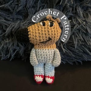 Puede incluir: Juguete amigurumi de crochet de un personaje de dibujos animados con pelaje marrón, ropa azul y zapatos rojos. El juguete está sentado sobre una superficie azul peluda. El texto "Crochet Pattern" está escrito en un círculo alrededor del juguete.