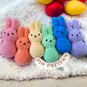 Può includere: Sei peluche di coniglietti all'uncinetto in un arcobaleno di colori, rosa, arancio, giallo, verde, blu e viola. I coniglietti sono disposti a semicerchio su una coperta bianca e soffice. Il testo "PDF PATTERN" è visibile nell'angolo in basso a destra dell'immagine.