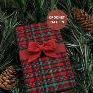 Op de afbeelding: Een ingepakte geschenkdoos met een rood-groen ruitpatroon, versierd met een gehaakte rode strik. De doos is verpakt met een rood lint. De tekst "CROCHET PATTERN" staat in een cirkel rechtsboven. De achtergrond bevat dennennaalden en dennenappels.