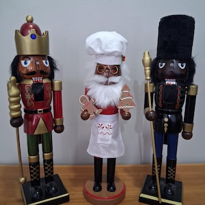 Può includere: Tre schiaccianoci decorativi in legno. Uno indossa una corona e un'uniforme rossa e oro, un altro un cappello da chef e un grembiule, e il terzo un cappello nero alto e un'uniforme scura. Ogni schiaccianoci ha un design unico e poggia su una base quadrata.