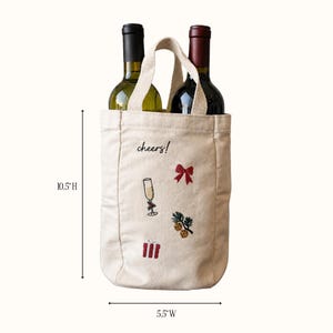 May include: A beige canvas wine tote bag with two handles, holding two wine bottles. The bag features embroidered designs of a champagne flute, a red bow, a gift box, and pine branches with the text cheers!. It measures 10.5 inches high by 5.5 inches wide.