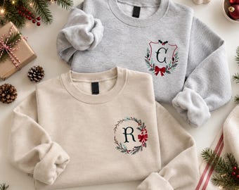 May include: A light gray sweatshirt featuring a crest with the letter C and a beige sweatshirt featuring a wreath with the letter R. Both garments have long sleeves, crew necklines, and embroidered holiday designs with red bows and greenery.