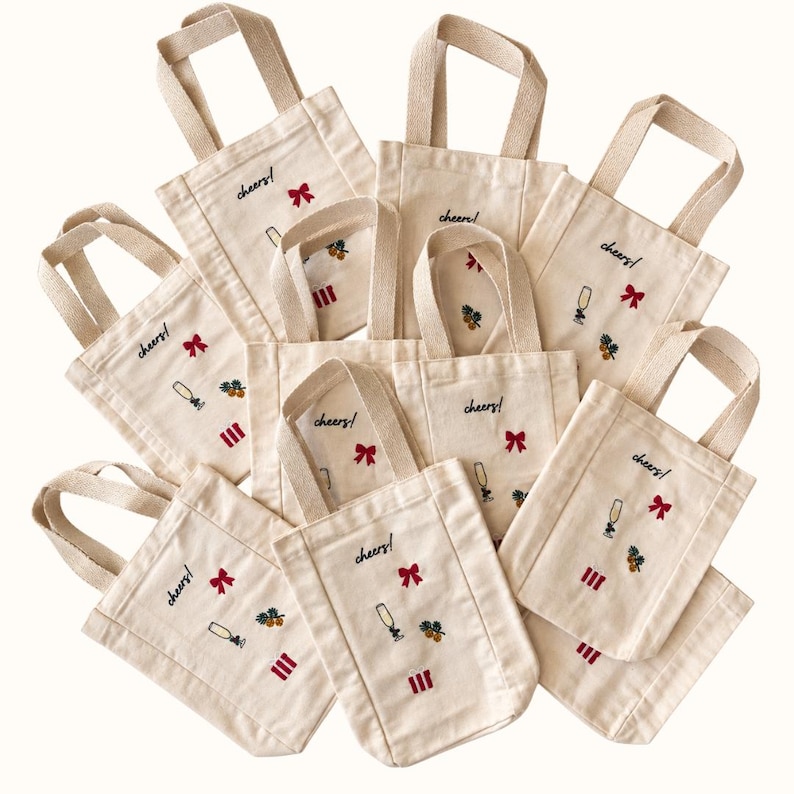 May include: A collection of off-white canvas tote bags, each featuring embroidered designs of a champagne flute, a red bow, a small cluster of pinecones, and three red vertical stripes, with the word cheers! embroidered in black script.