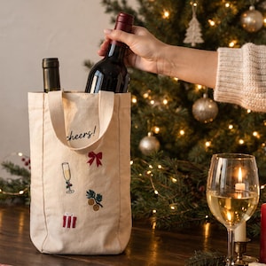 May include: A cream-colored canvas wine tote bag featuring the embroidered text cheers! and festive motifs including a red bow, a champagne flute, pine branches with oranges, and a gift box. The bag holds two wine bottles with dark glass.