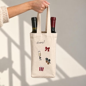 May include: A beige canvas wine tote bag with two handles, holding two wine bottles. The bag features embroidered designs including a red bow, a champagne flute, a pine branch with ornaments, a red gift box, and the word cheers in black script.