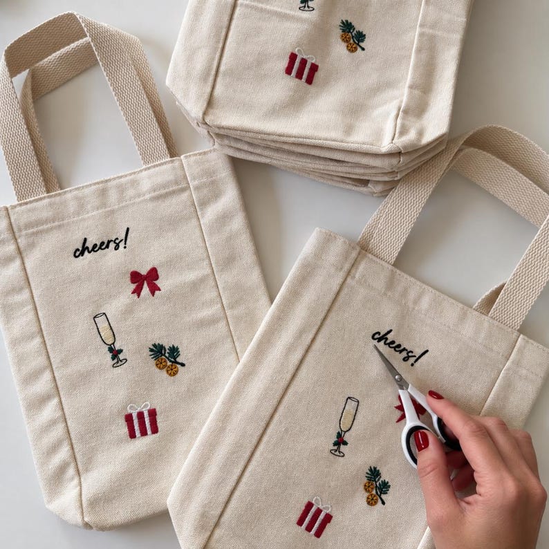 May include: Off-white canvas tote bags featuring embroidered designs of a champagne flute, a red bow, a gift box, and pine sprigs with berries. The text "cheers!" is embroidered in black thread above the designs on each bag.