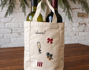 May include: A beige canvas wine tote bag with two handles, holding two wine bottles. The bag features embroidered designs including a red bow, a champagne flute, a gift box, and pine branches with orange slices, along with the text cheers! in black.