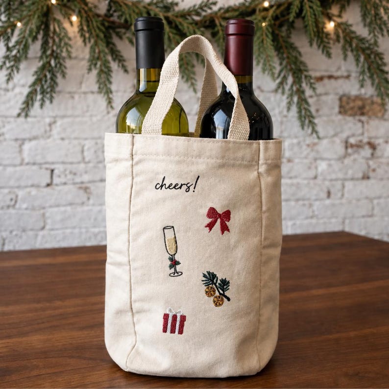 May include: A beige canvas wine tote bag with two handles, holding two wine bottles. The bag features embroidered designs including a red bow, a champagne flute, a gift box, and pine branches with orange slices, along with the text cheers! in black.