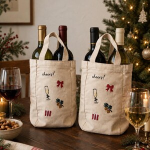 May include: Two off-white canvas wine bottle bags with handles, each embroidered with the word cheers, a champagne flute, a red bow, a pine sprig, and a gift box. Each bag holds a wine bottle, one with a gold foil and one with a black foil.