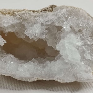 Peut inclure: Une géode blanche avec une grande ouverture révélant un intérieur tapissé de cristaux. La géode est une formation rocheuse naturelle avec un extérieur rugueux et un intérieur lisse et blanc.