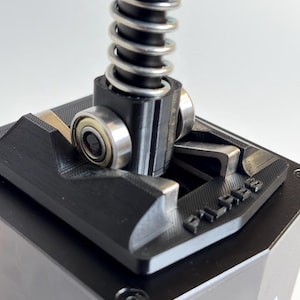 Puede incluir: Primer plano de un componente mecánico negro y plateado. Un resorte plateado está conectado a una pieza cilíndrica negra con dos rodamientos. El componente está montado sobre una base negra con la palabra "PILABS" en relieve.