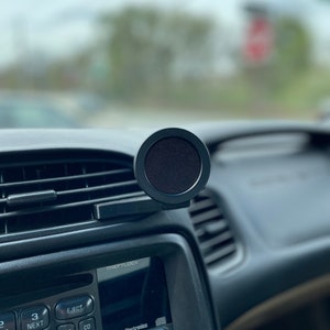 Puede incluir: Una radio de coche negra con botones etiquetados "Eject", "AM", "CD", "FWD", "FM", "AUX", "RECALL", "FADE", "BAL", "BASS", "TREB", "AUTO" y "TONE". La radio tiene una pantalla negra con el texto "THEFTLOCK" y "Delco Electronics" mostrado. Un soporte de teléfono negro para ventilación de coche está fijado a la ventilación por encima de la radio.