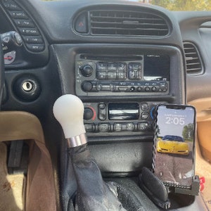 May include: A black car interior with a white gear shift knob, a black radio, and a black phone holder with a smartphone displaying the time 2:05. The phone screen shows a yellow car.