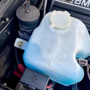 May include: A close-up of a blue windshield washer fluid reservoir with a white cap and a black plastic cap on a black engine compartment. The reservoir has the text "1.5L" printed on the side.