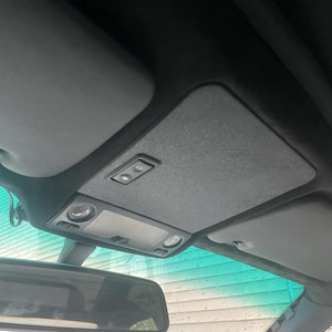 Puede incluir: Vista interior del coche que muestra la consola superior con una lámpara, parasoles y espejo retrovisor. La consola es gris con dos botones pequeños. Los parasoles son grises y el parabrisas tiene un tinte verde.