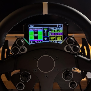 Könnte beinhalten: Schwarzes Rennlenkrad mit digitaler Anzeige, die Leistungsdaten anzeigt. Das Lenkrad hat mehrere Knöpfe und Drehregler mit einem weißen Streifen oben. Die Anzeige zeigt Rundenzeiten, Geschwindigkeit und andere Messwerte.