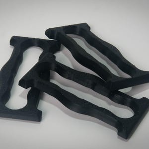 May include: Four black, 3D-printed handles are arranged on a white surface. Each handle has a rectangular base, a curved grip, and a top section with a similar rectangular shape. The handles appear to be made of a matte material.