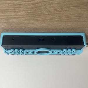 Puede incluir: Un dispositivo informático negro con una carcasa protectora azul claro. El dispositivo tiene un puerto USB y un puerto de alimentación en la parte posterior. La carcasa tiene un diseño decorativo en la parte inferior.