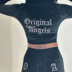 May include: Black velvet tracksuit with a cropped hoodie and matching pants. The hoodie features a rhinestone design of wings and the text "Original Angels". The pants have a rhinestone "O" on the left back pocket and a rhinestone "A" on the right back pocket.