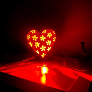 Könnte beinhalten: Eine rote herzförmige Lampe mit einem Blumenmuster und einem gelben Schein. Die Lampe steht auf einer weißen Oberfläche und wirft ein rotes Licht.
