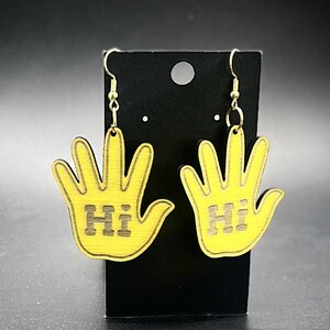 Puede incluir: Un par de pendientes amarillos con forma de mano con la palabra "Hi" recortada en el centro de cada mano. Los pendientes cuelgan de una tarjeta de exhibición negra con ganchos dorados.