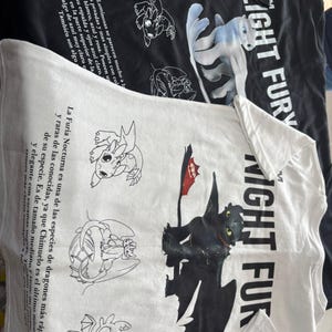 May include: Two t-shirts, one black and one white, featuring dragon graphics. The black shirt has a white dragon and the text "LIGHT FURY." The white shirt displays a black dragon with a red flag and the text "NIGHT FURY."