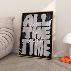 May include: Framed art print with the text "ALL GOD IS THE GOOD TIME" in white, bold, handwritten-style lettering on a black background. The print is in a gold-colored frame and leans against a white wall.