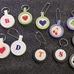May include: A collection of 12 keychains with different embroidered designs. The keychains are made of white, green, purple, and black plastic with metal rings. The designs include a rainbow heart, a red heart, a strawberry, a blue heart, a cherry, a four-leaf clover, a ladybug, a butterfly, and the letters B, D, T, and S.