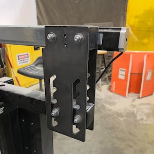May include: A close-up of a black metal bracket with multiple bolt connections, attached to a silver metal beam. The bracket has a series of cut-out shapes and is mounted on a larger black structure. The background includes a yellow cabinet and orange boxes.