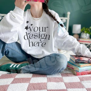 May include: White sweatshirt with the text "your design here" in black. The person is wearing blue jeans and green sneakers. A stack of books is visible on the bed. The bedspread has a pink and white checkered pattern.